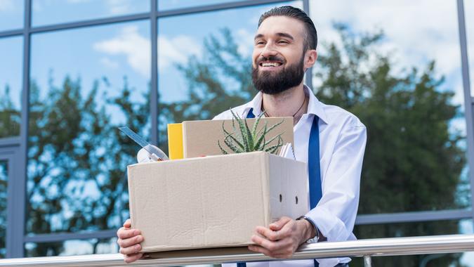 happy businessman with cardboard box with office supplies in hands standing outside office building, quitting job concept.