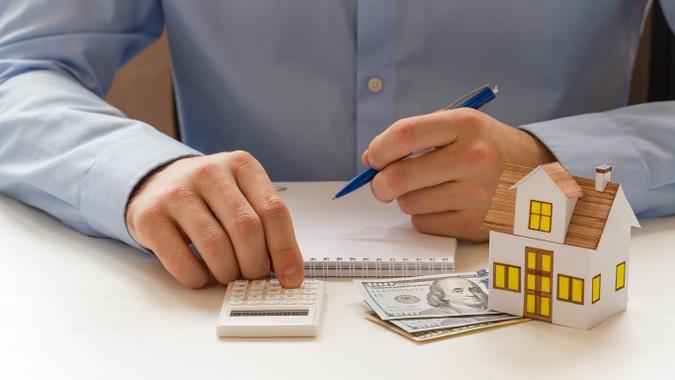 man calculating finances next to toy house sitting on fan of $100 cash money