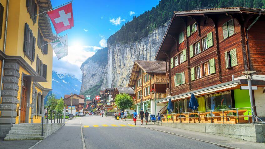 Bernese Oberland, Europe, How Much to Tip When Traveling to These 25 Countries, Principal street of Lauterbrunnen with shops, Switzerland, hotels, swiss flags and stunning Staubbach waterfall in background, terraces