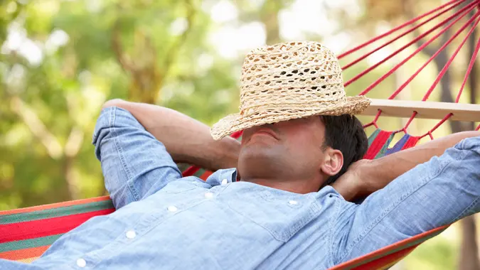 A man laying in a hammock with his arms behind his head and a sun hat over his face.