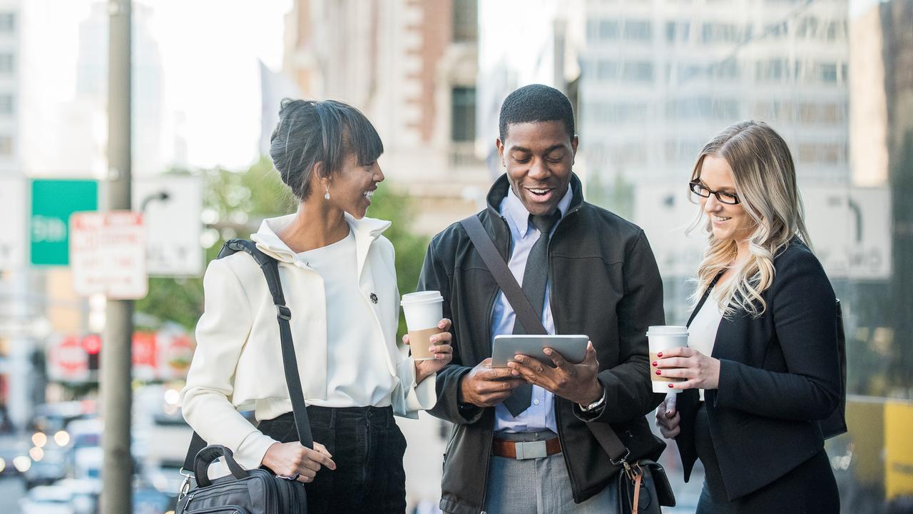 A young African American male and female and a young blonde caucasian female business associates or colleagues, on a coffee break, engaged in light-hearted conversation involving content on the digital tablet on the side of a city street.