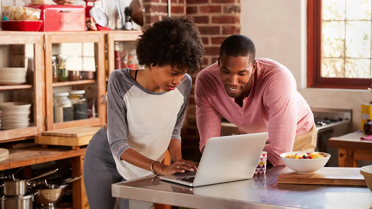 Two young adults sit together at a table, smiling and looking at a laptop screen in a well-lit room.