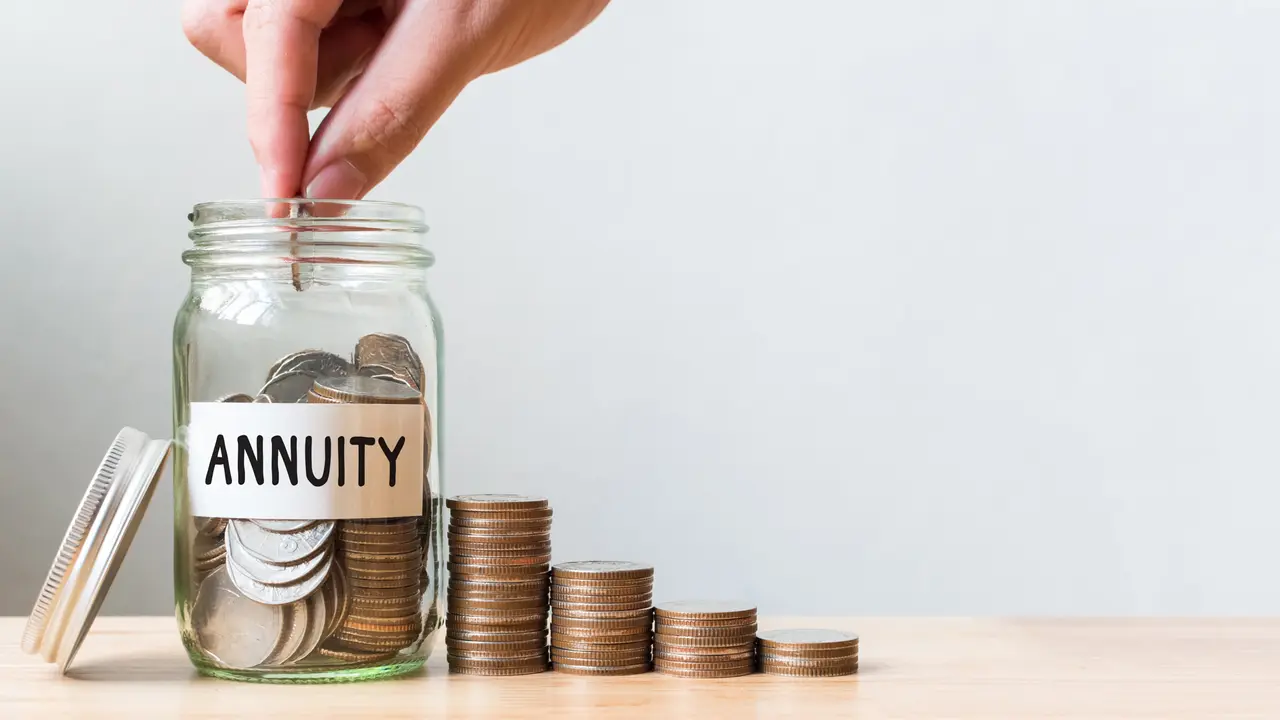 Hand placing coin into a jar labeled "Annuity" beside stacks of coins increasing in height on a wooden surface.