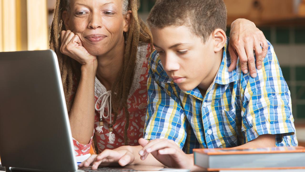 Mother overlooks as her teenaged son uses his laptop to study in their kitchen.