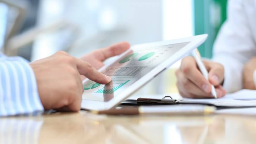 one man writing on a piece of paper and another man looking at charts on his ipad tablet