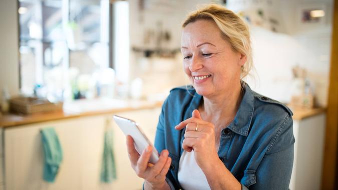 Beautiful senior woman at home in her kitchen holding smart phone, writing or reading something, smiling.