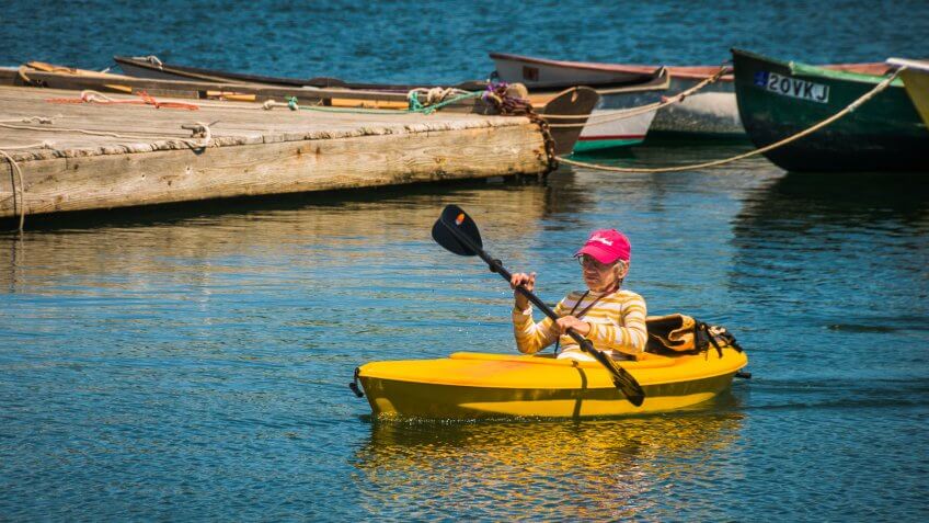 Round Pond, Maine, USA-June 14, 2017- A mature woman paddles her yellow kayak past a pier and boats to reach her take out spot after an envigorating solo paddle on Round Pond off Muscongus Bay.