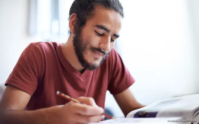 A man Sitting at a desk and looking at a textbook while holding a pencil.