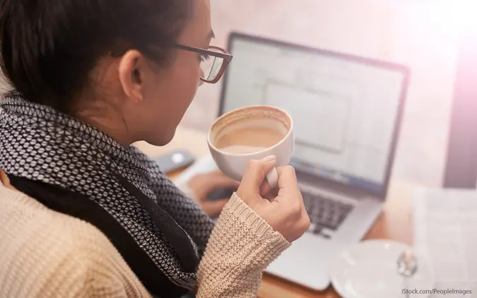 Woman in glasses and beige sweater looks at loan info on laptop while holding a cup of tea