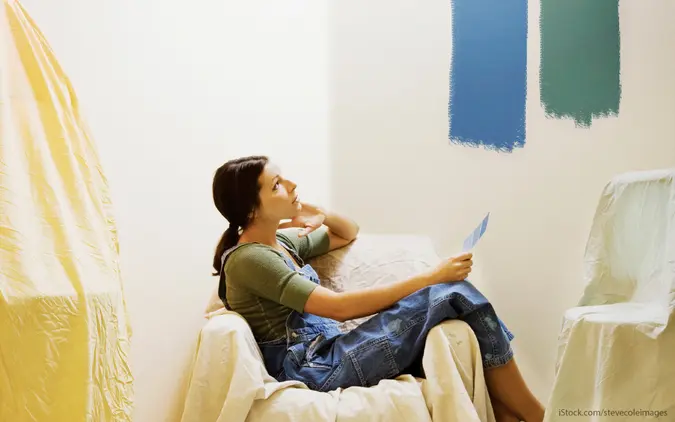 Surrounded by draped furniture, a woman ponders two paint colors, holding swatches and staring at the wall in thought.