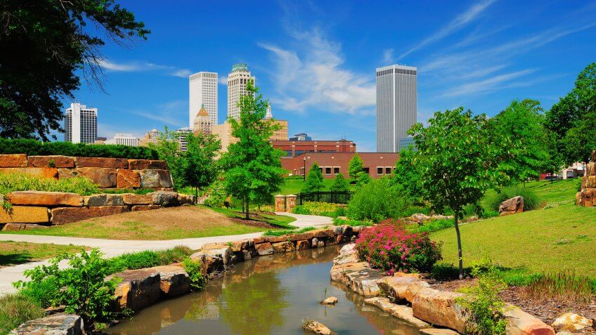 Tulsa downtown skyline from a park with trees, grass, rocks, and a stream in the foreground.