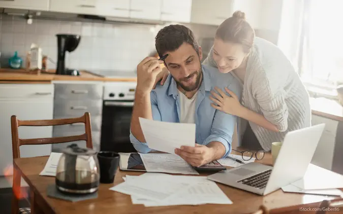 A couple evaluates their finances at the kitchen table, with a laptop, paperwork, and coffee on the table.