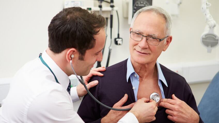Doctor Examining Senior Male Patient In Hospital Listening To Heartbeat Using Stethoscope.