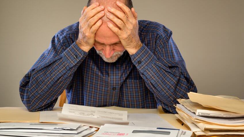 A senior adult man sitting at a table or desk stacked with papers and envelopes looking down with his hands on his head.