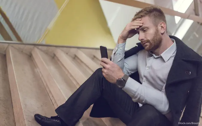 Stressed man sits on steps, anxiously looking at his phone—he may have just fallen victim to a scam.