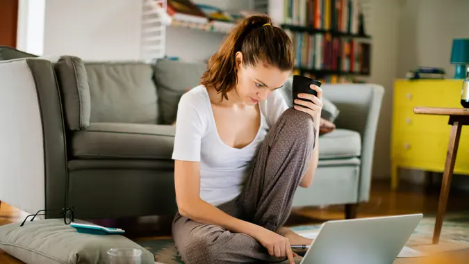 A woman thoughtfully reviews her finances on her laptop and phone while sitting on her cozy living room floor.