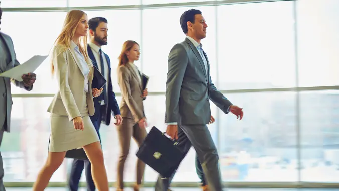 A wealthy businessman or CEO leading his employees down a hallway inside a building or workplace.