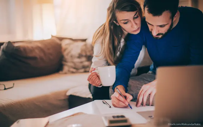 A couple sits on a sofa, intently reviewing financial documents on their coffee table with a laptop and calculator.