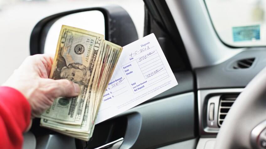 A man in his car is holding five hundred dollars in US twenties and a bank deposit slip while waiting in line for a bank drive through teller window.