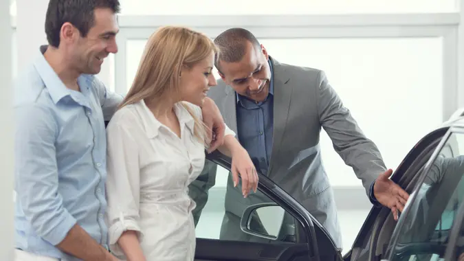 A happy couple talks with a car salesperson at a dealership while considering a new vehicle.