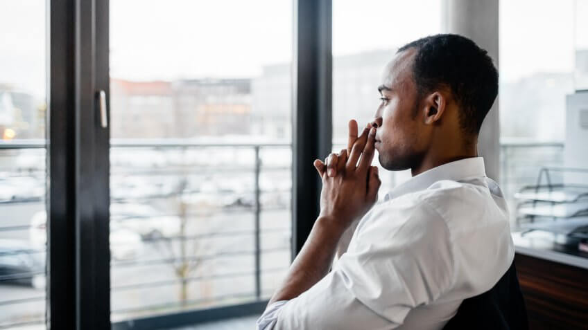 Pensive and serious young black businessman sitting in a chair in his office and looking through a window away.