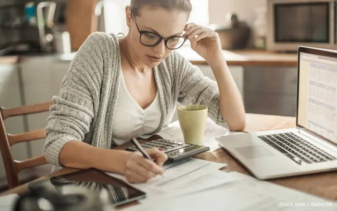 A woman wearing glasses reviews financial paperwork at her desk with a laptop open in front of her.