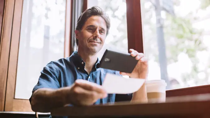 A mature smiling man in his mid 40's takes a picture with his smart phone of a check or paycheck for digital electronic depositing, also known as "Remote Deposit Capture".
