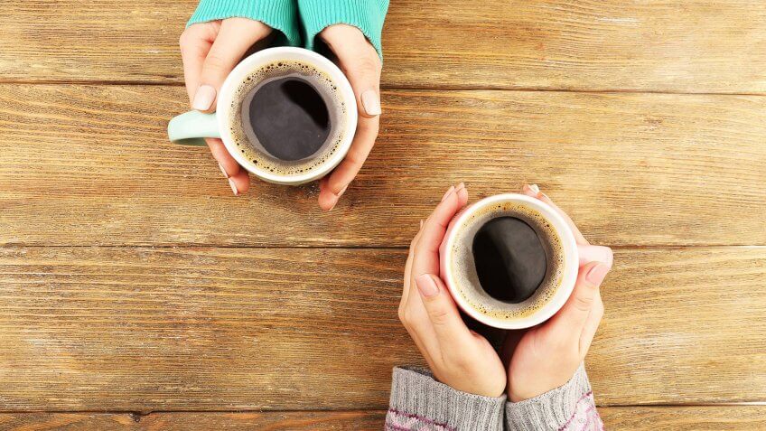 Female hands holding cups of coffee on rustic wooden table background.