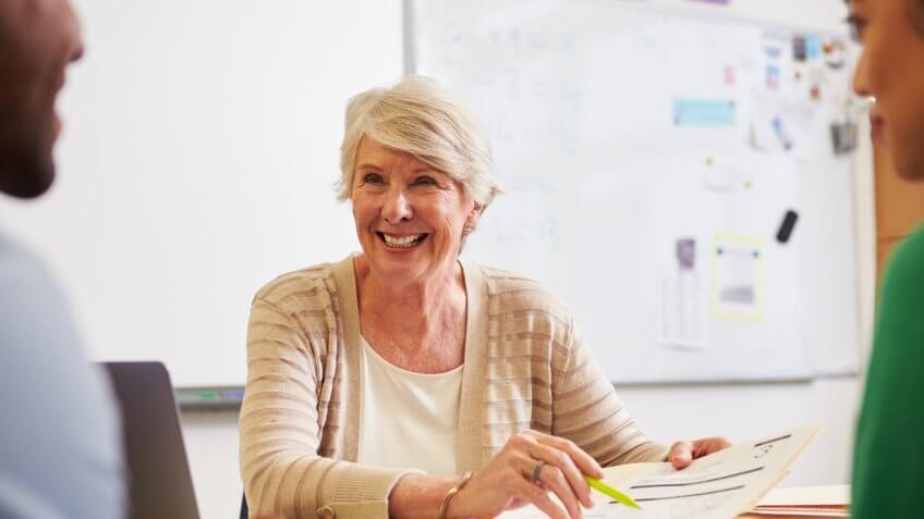 Senior teacher at desk talking to adult education students.