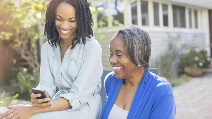 Smiling mother and daughter text messaging with cell phone on patio.