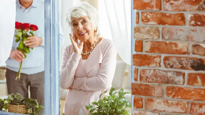 Old happy woman looking through the window with small green plants.