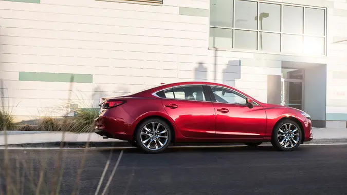 A red Mazda6 Sport sits parked on the street outside a white building with the sun shining on it.
