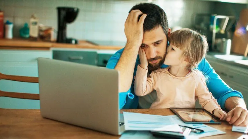 daughter kissing father on the cheek while father looks upset at his documents and laptop