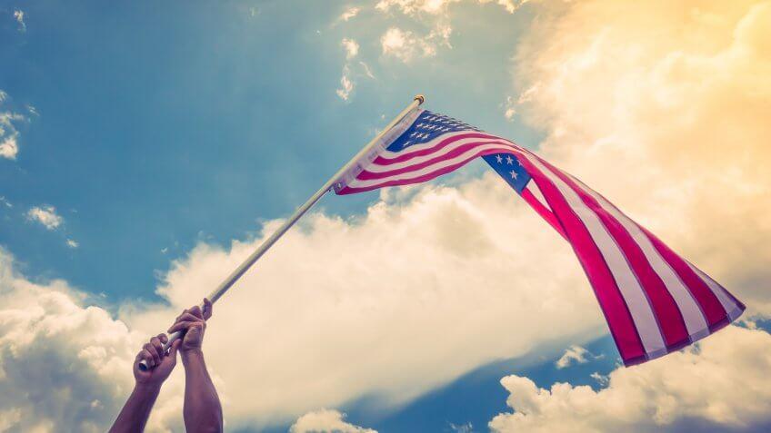 American flag with stars and stripes hold with hands against blue sky.