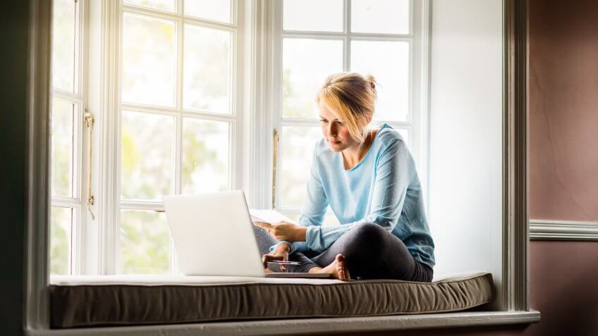 A full length photo of woman using laptop while holding documents at home.