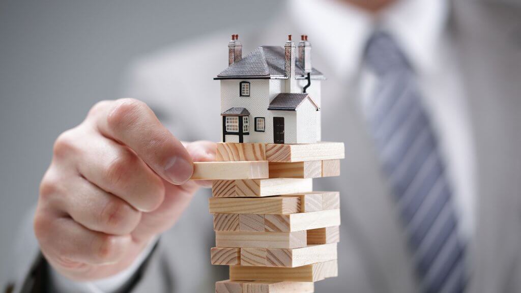 The housing market is unpredictable. tiny model house on top of jenga stack