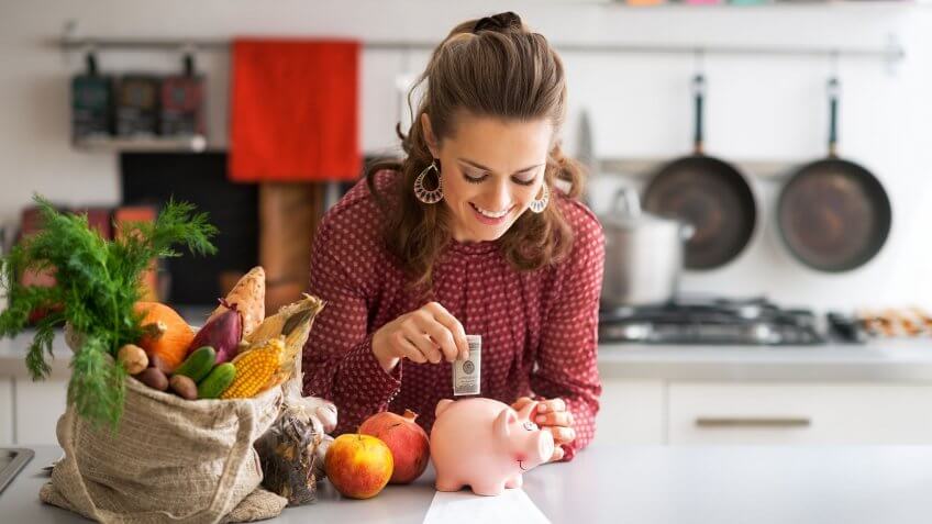 woman in kitchen putting 100 dollars in a piggy bank