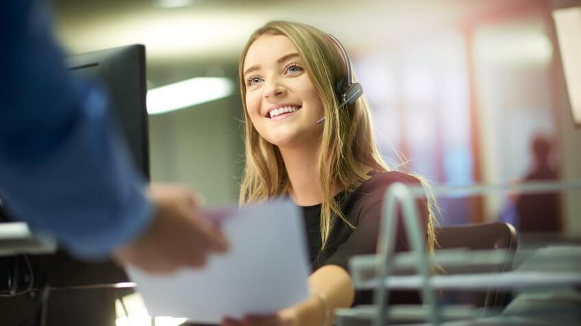 a young woman sits at her desk and is handed some paperwork by a colleague out of shot.