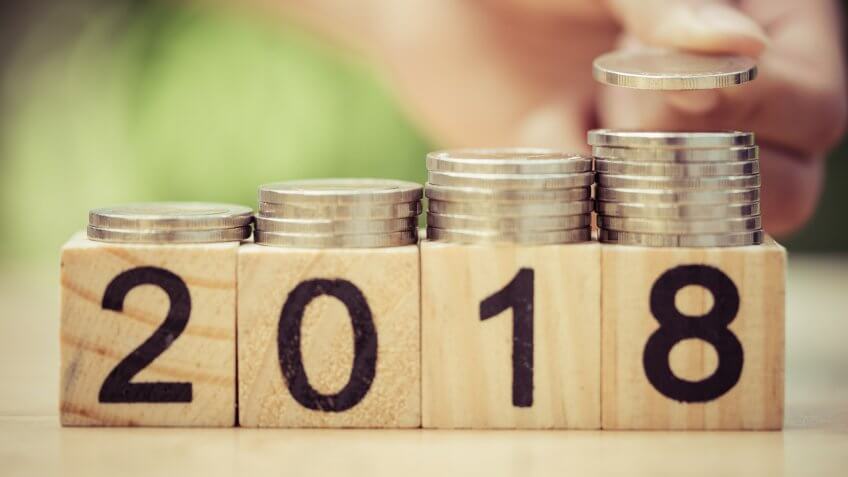 hand putting coins to stack of coins on wooden block with text 2018.