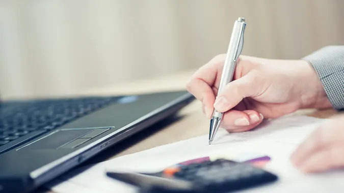 Close-up image of a person writing on a piece of paper with a laptop and calculator next to them.
