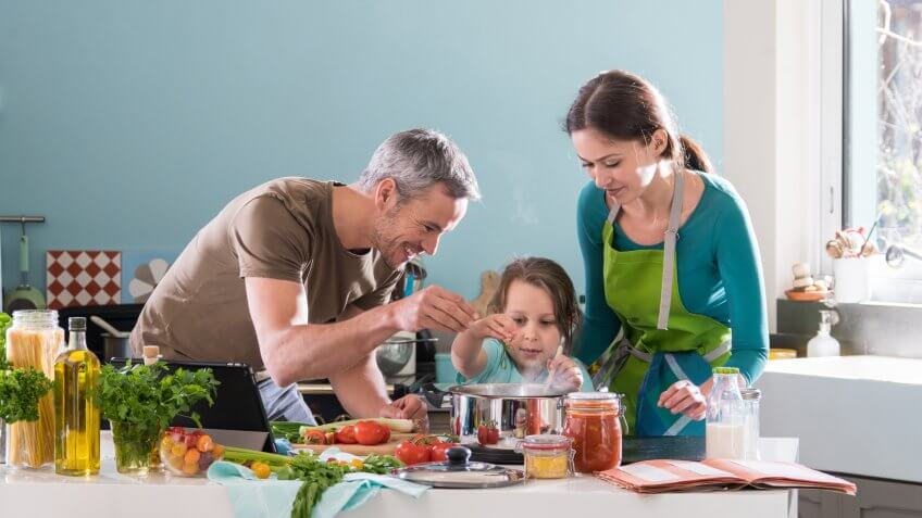 Dad, Mom and their little daughter cooking bolognese sauce for spaghetti in the kitchen, there is steam escaping from the pan on cooking plate.