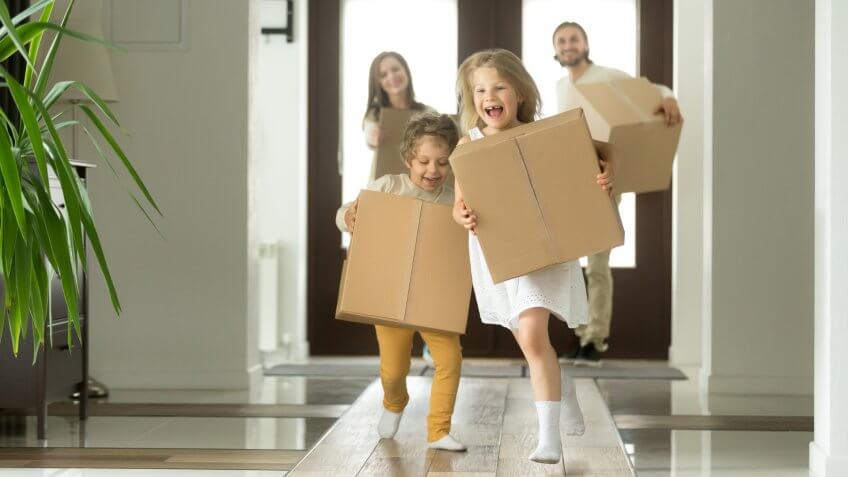 Happy family with kids bought new home, excited children funny girl and boy holding boxes running into big modern house, helping parents with belongings, moving day concept, mortgage and relocation.