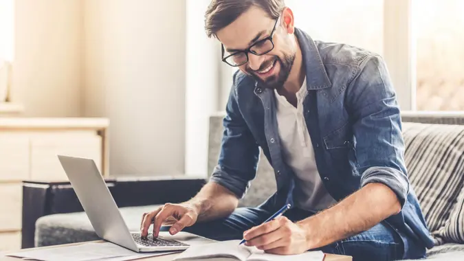 Handsome businessman in eyeglasses is making notes and smiling while working with a laptop at home.