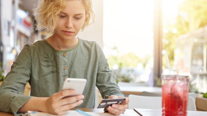 mujer en blusa sentada en un café sosteniendo un teléfono móvil y una tarjeta de plástico registrándose en el sitio web