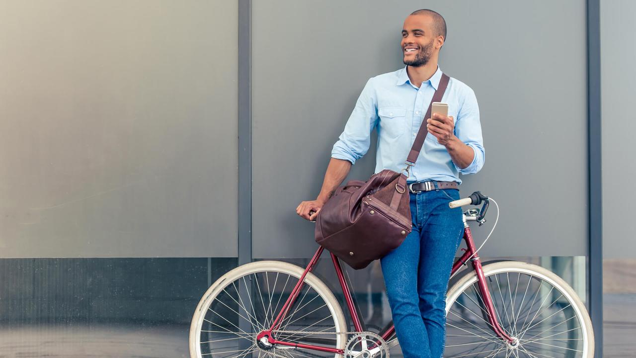 Full length portrait of handsome young Afro American man in casual clothes using phone, looking away and smiling while leaning on his bike, standing outdoors.