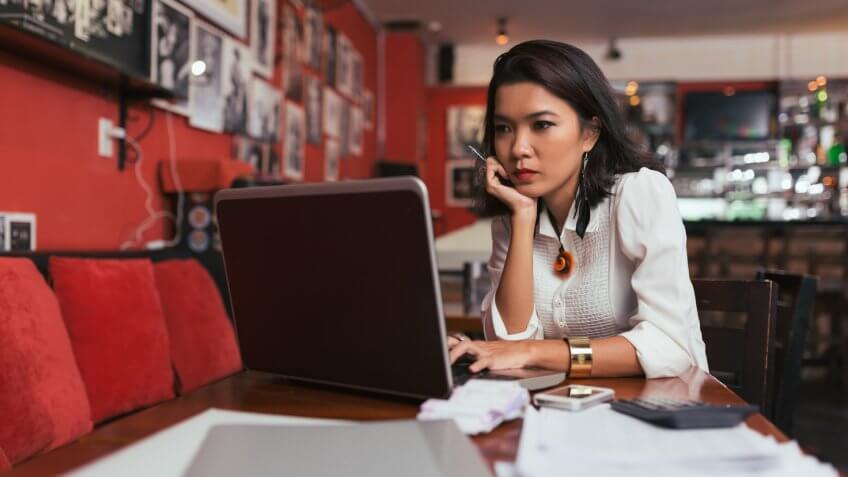 Pensive Asian bar owner using laptop to pay bills online.