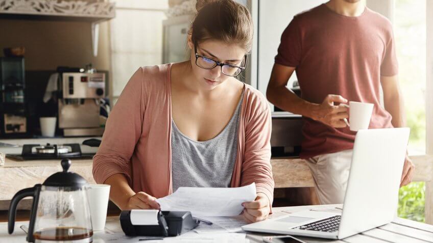 woman holding paper calculating numbers with man drinking coffee behind her