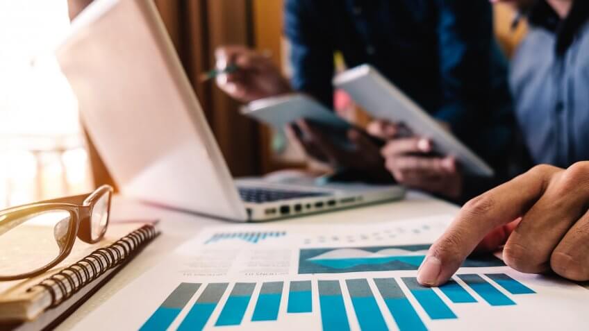 close up of man pointing to printed chart graph with people working in the back