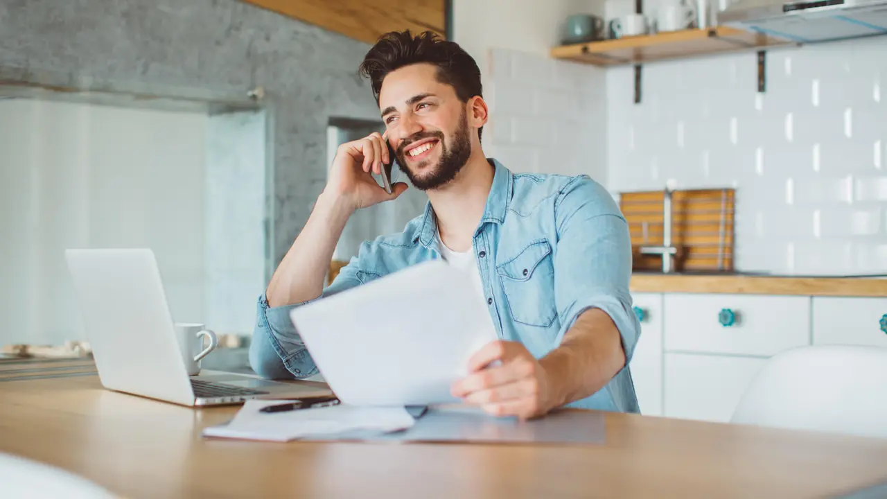 Young man sitting at the table in a kitchen, working on laptop and making phone call.