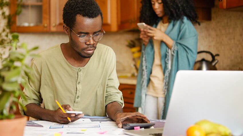 Man calculating finances with woman on her phone behind him